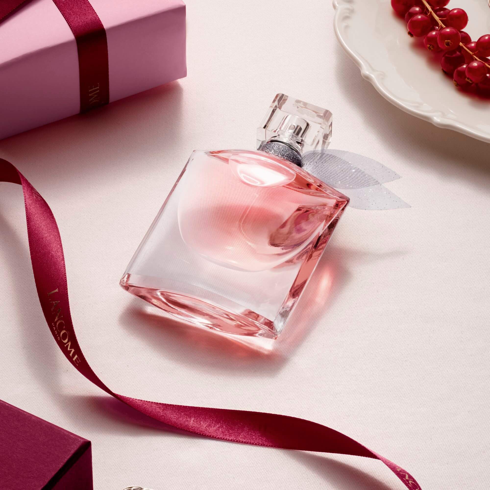 A pink Lancome perfume bottle styled on a light surface with a gift box, ribbon, and red currants.