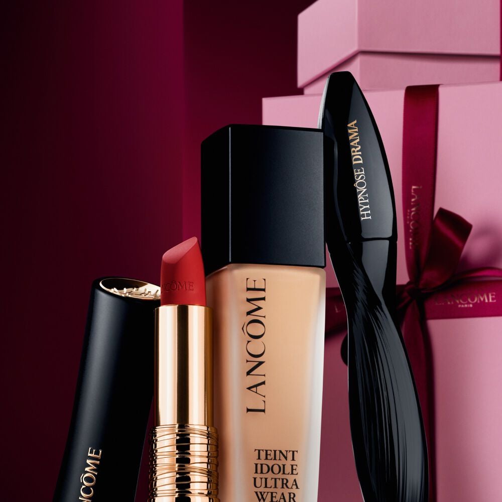 An elegant still life of Lancome's L'Absolu Rouge matte lipstick, Teint Idole Ultra Wear foundation, and Hypnose Drama mascara set against a luxurious dark red holiday background.
