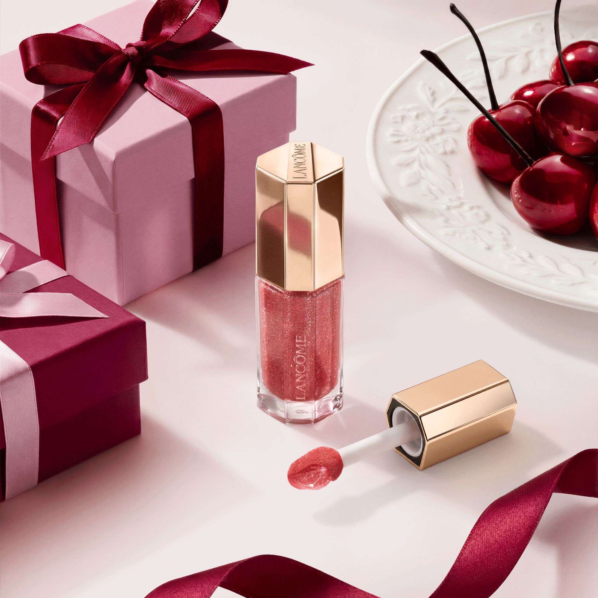 A flat-lay arrangement of Lancome makeup products, featuring mascara and lip gloss, alongside pink gift boxes, satin ribbons, and a sprig of red currants.