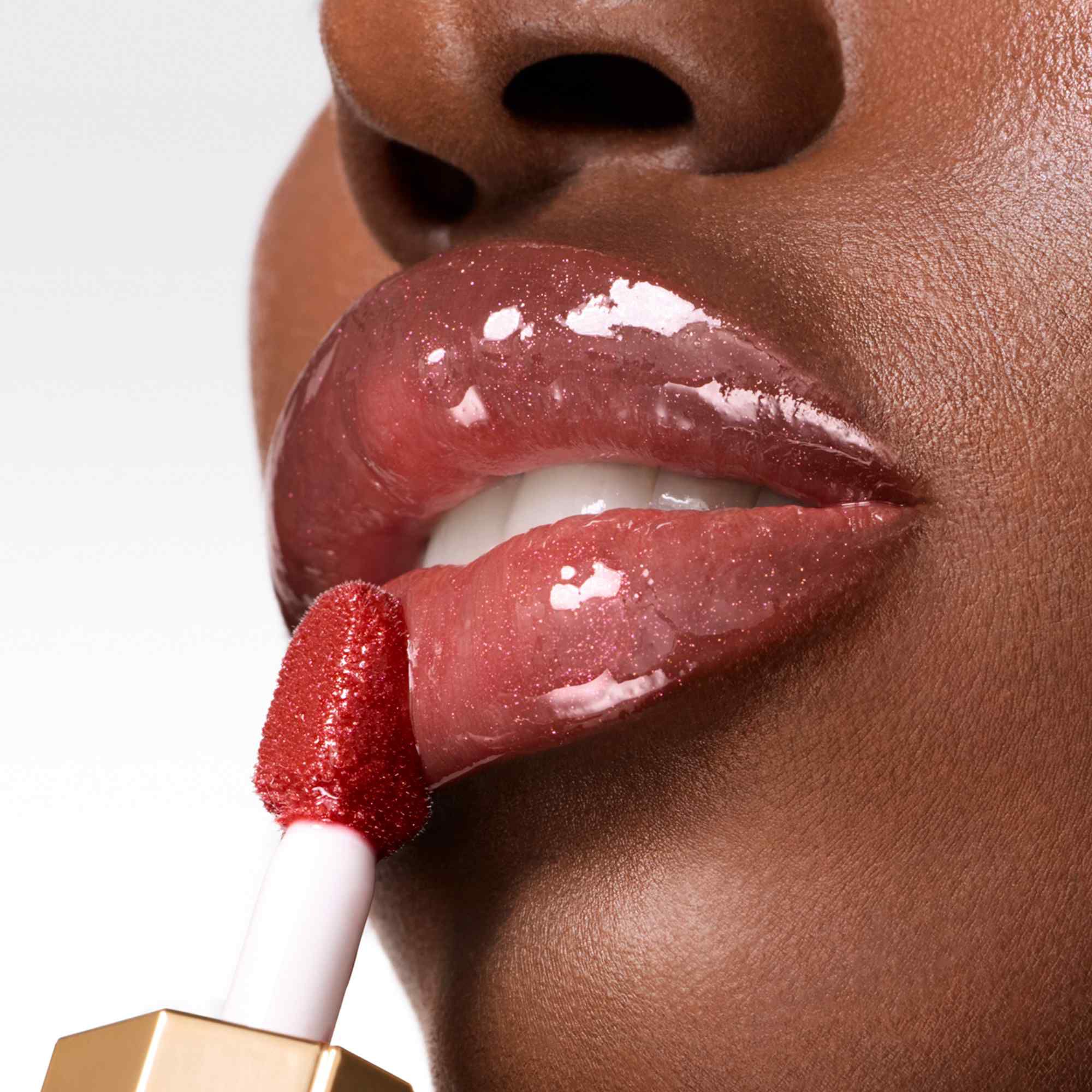 A close-up shot of a lip gloss wand applying a shimmering, glossy berry-toned product to a person's lips.