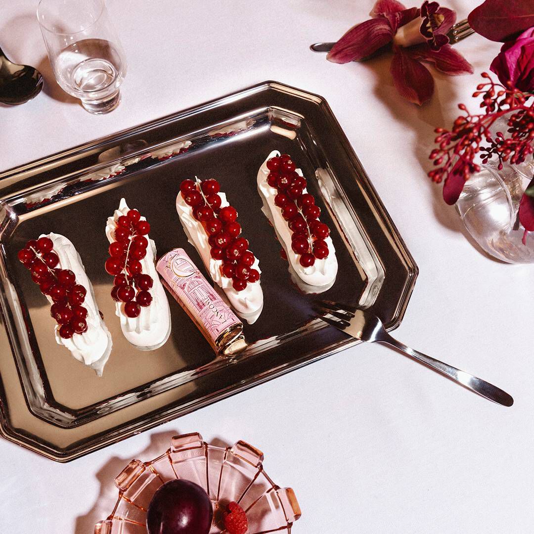 A Lancome lipstick nestled among four cream-topped desserts garnished with red currants on a silver tray.